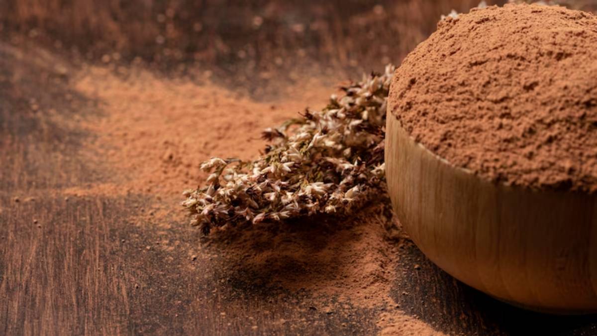 A wooden bowl filled with brown powder and scattered on a rustic surface, alongside dried flowers.