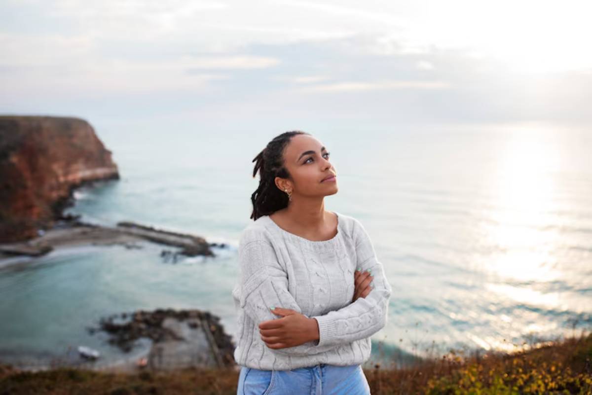 A person in a cozy gray sweater stands confidently by the seaside, with cliffs and soft waves in the background under a pastel sky.