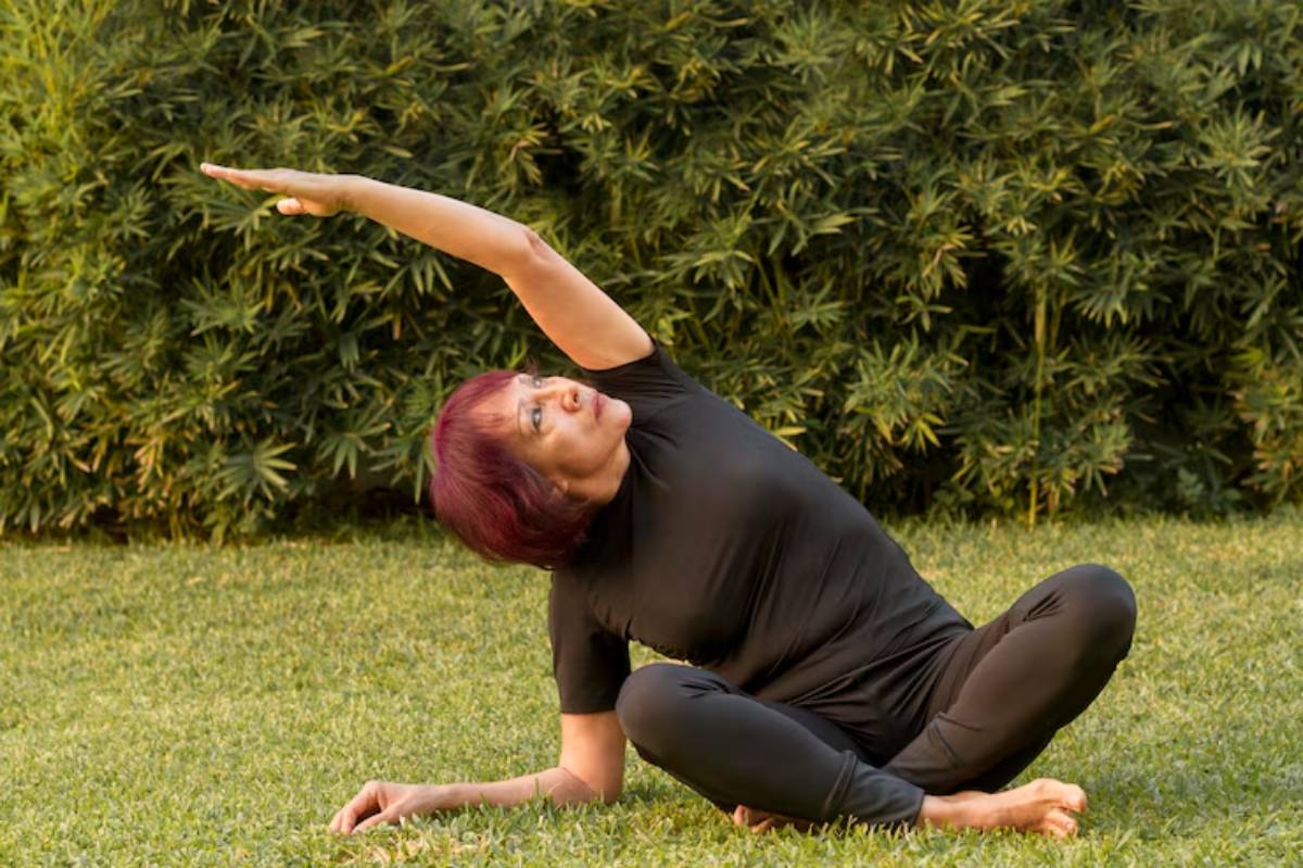 A person in black activewear performs a yoga pose on grass, stretching one arm overhead against a backdrop of lush greenery.