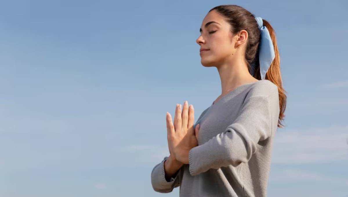 A woman with a ponytail and light sweater stands in a serene outdoor setting, practicing yoga with hands together in contemplation.