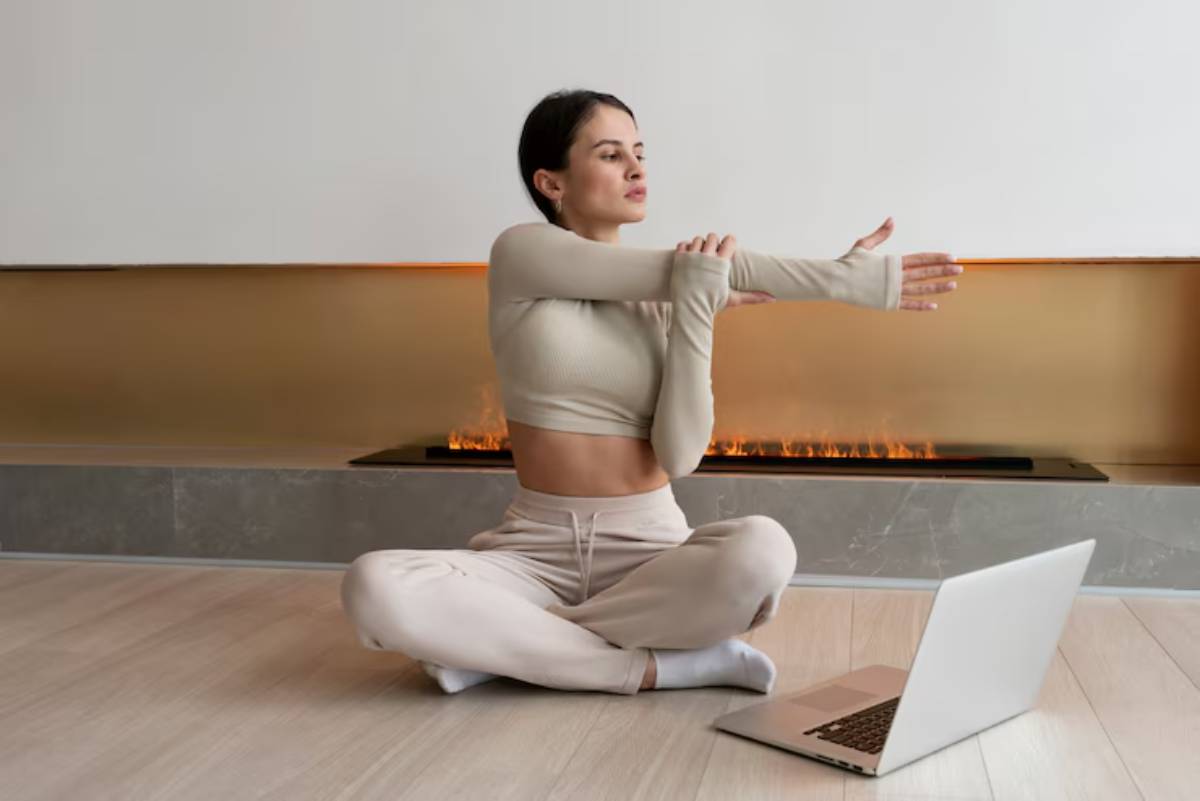A person in a light-colored outfit stretches beside a laptop, with a modern fireplace in the background.