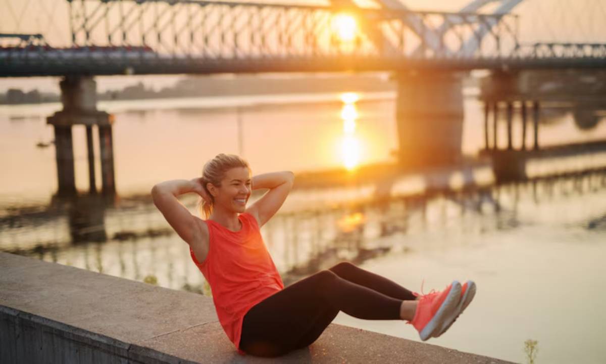 A person in a red tank top performs sit-ups by a riverside at sunset, with a bridge silhouetted in the background.