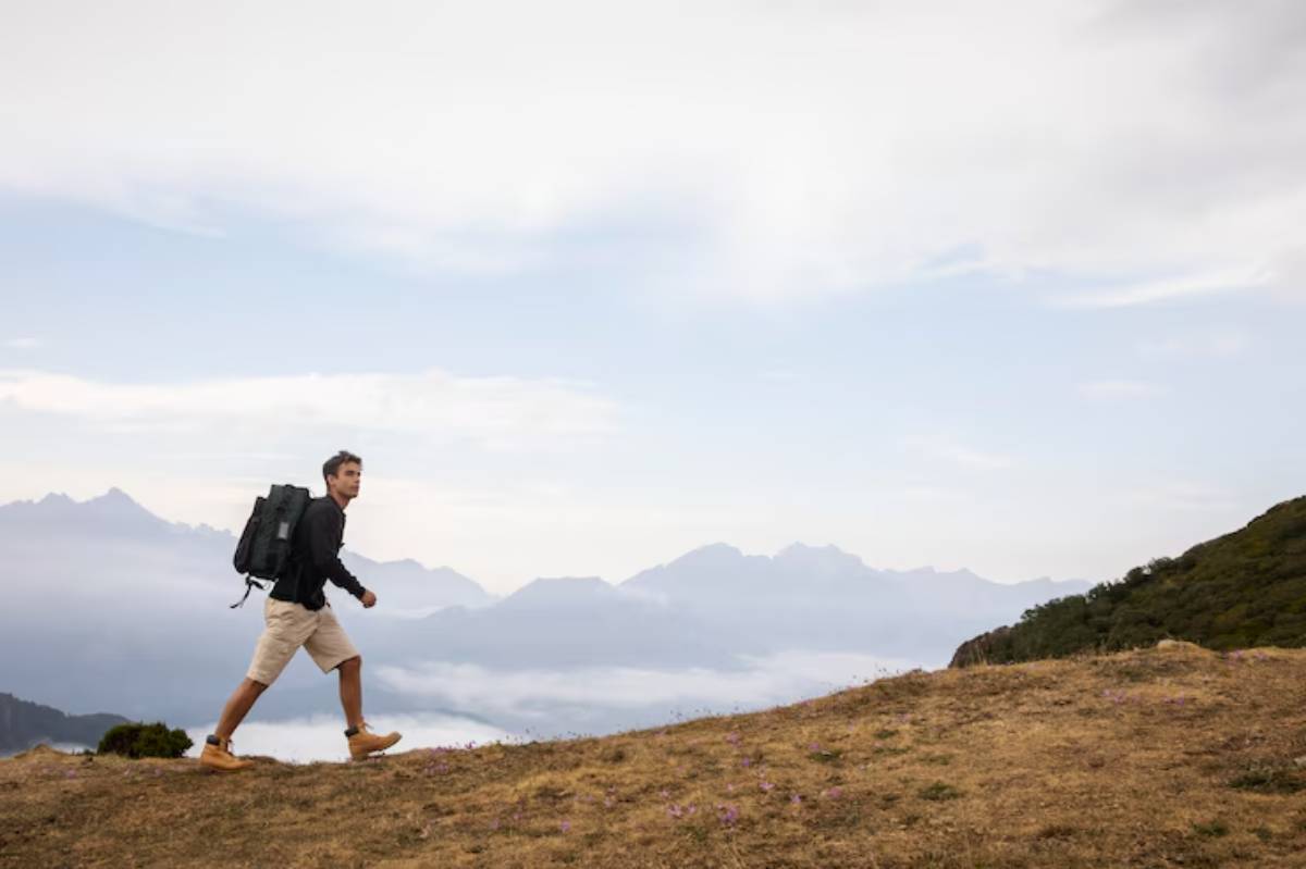 A hiker with a large backpack walks on a grassy hill, surrounded by misty mountains beneath a cloudy sky.