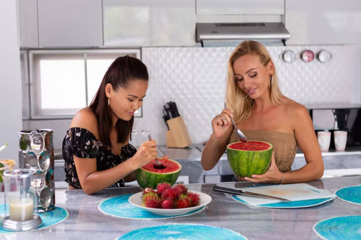 Two women enjoy scooping watermelon in a modern kitchen, surrounded by colorful platters and fresh fruit.