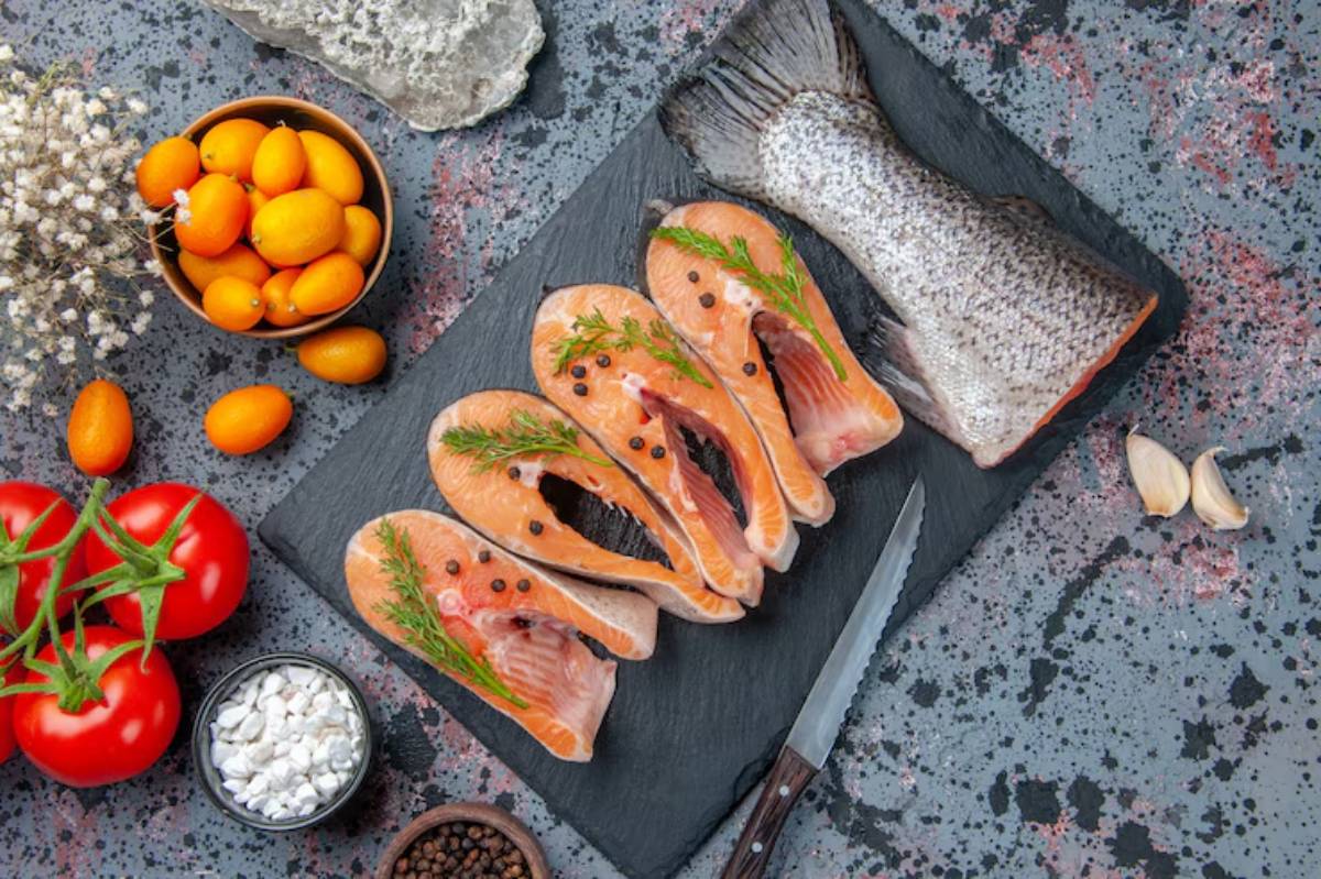A slate platter with salmon fillets garnished with herbs, whole fish, tomatoes, kumquats, garlic cloves, and seasoning.