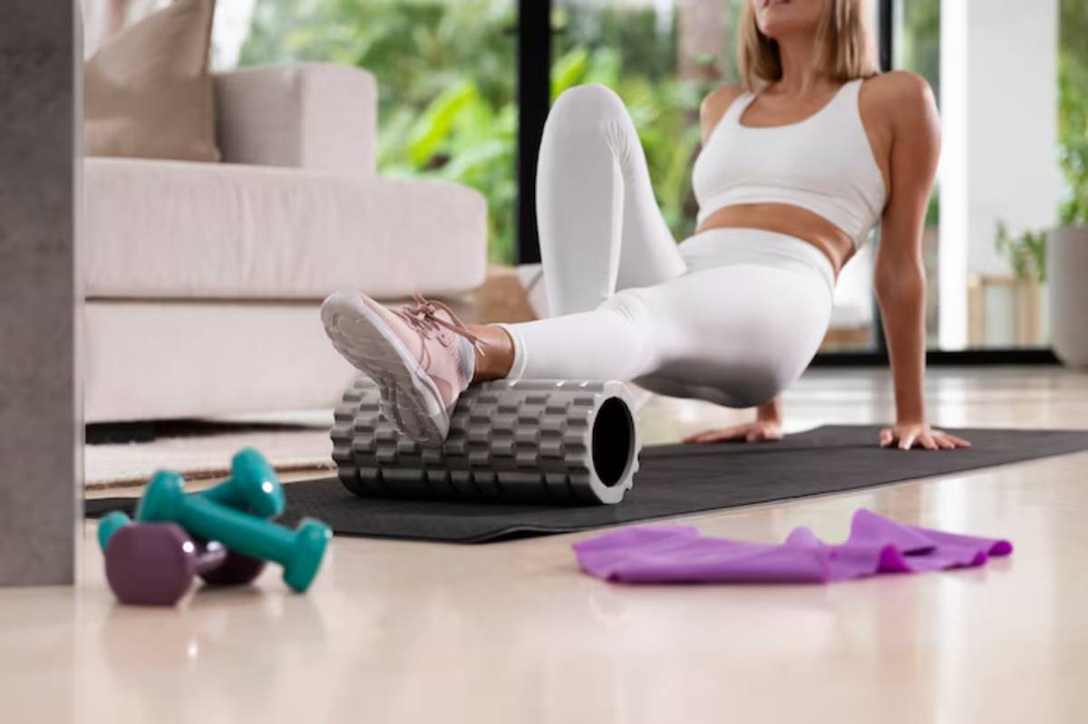 A woman in white athletic wear uses a foam roller on a yoga mat at home, surrounded by weights and a resistance band.