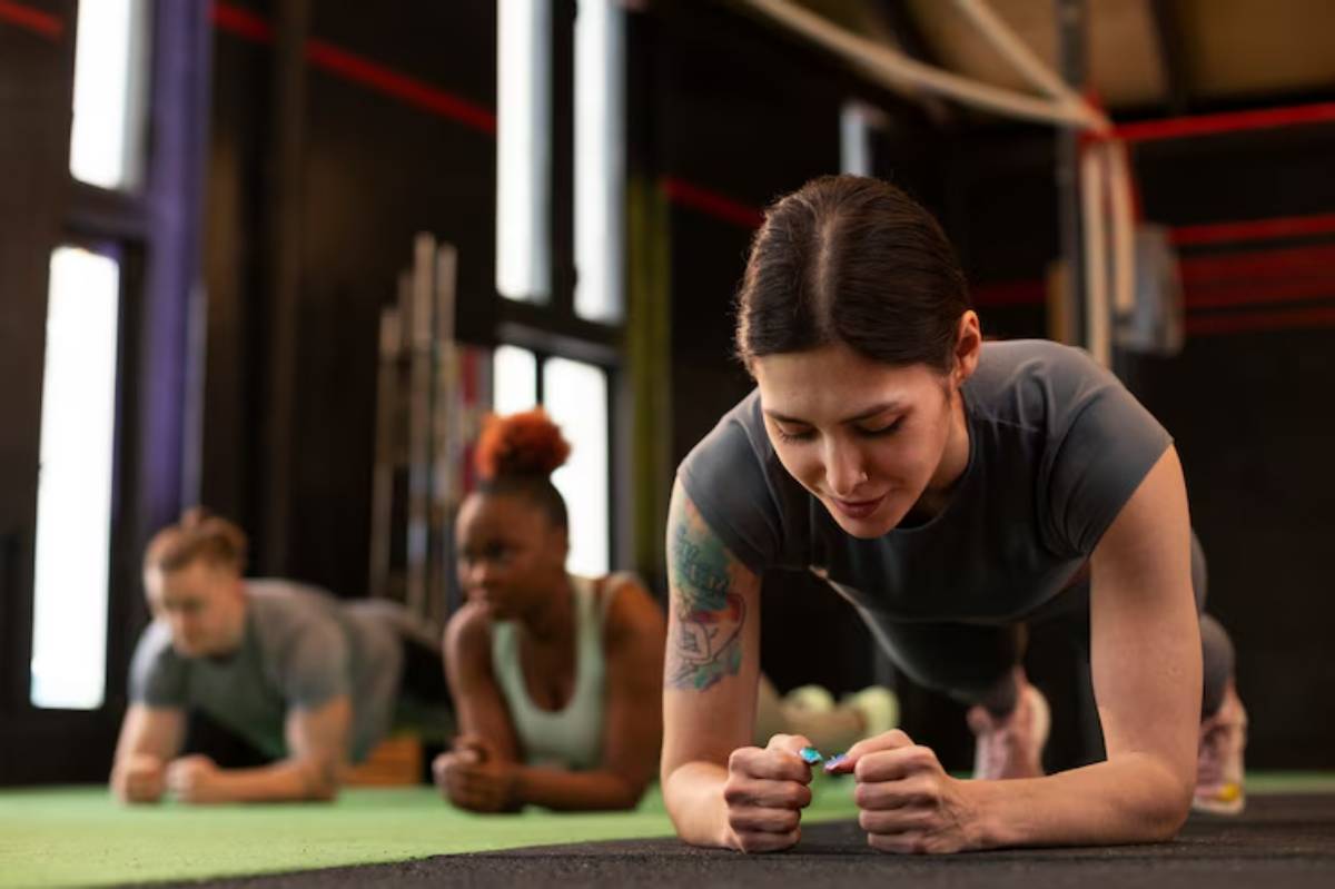 Three individuals perform plank exercises on a gym floor, surrounded by fitness equipment and bright natural light.