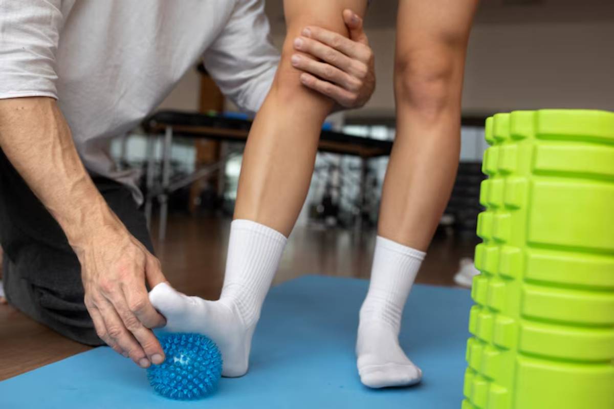 A therapist assists a patient in rolling their foot on a blue massage ball during a rehabilitation session in a gym setting.