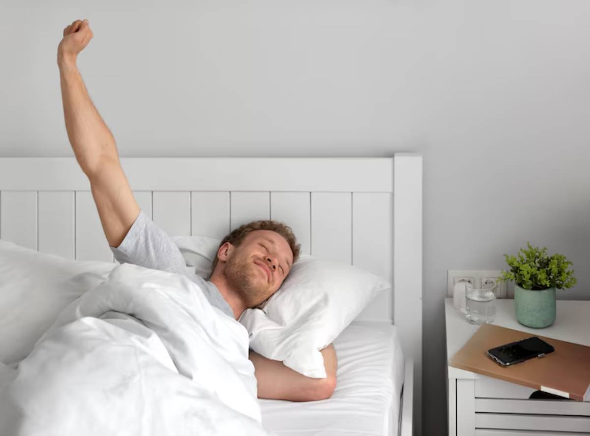 A person stretches in a bed, surrounded by white bedding, with a nightstand holding a phone and a small plant.