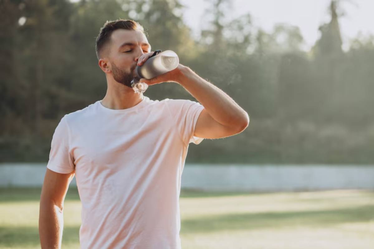 A person in a white t-shirt enjoys a refreshing drink outdoors, surrounded by greenery in warm sunlight.