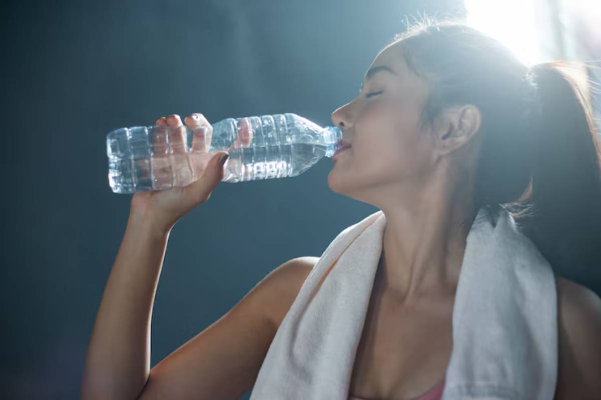 A fit individual takes a refreshing drink from a plastic water bottle, with a towel draped around their neck, against a moody backdrop.