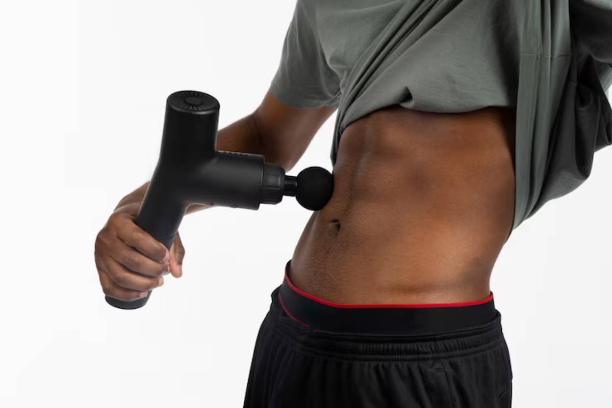A person using a handheld massage device on their abdomen, wearing a gray shirt and black shorts with a red waistband.