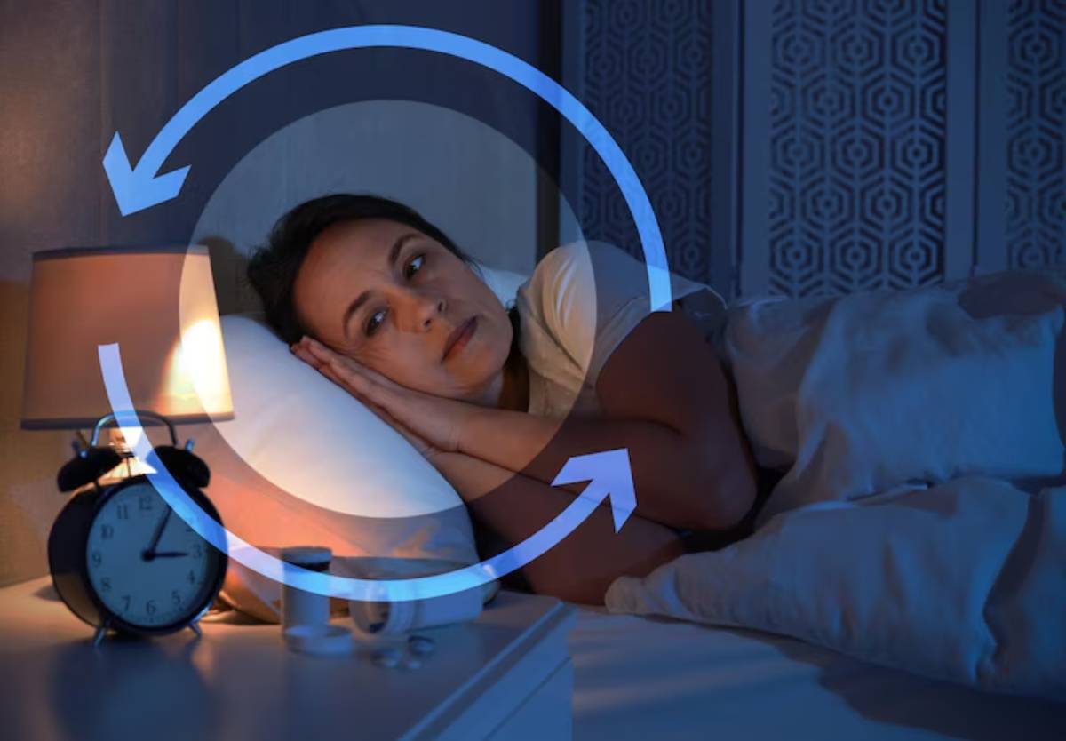 A person lays in bed under dim blue light, surrounded by pillows and a bedside table with a lamp and an alarm clock.