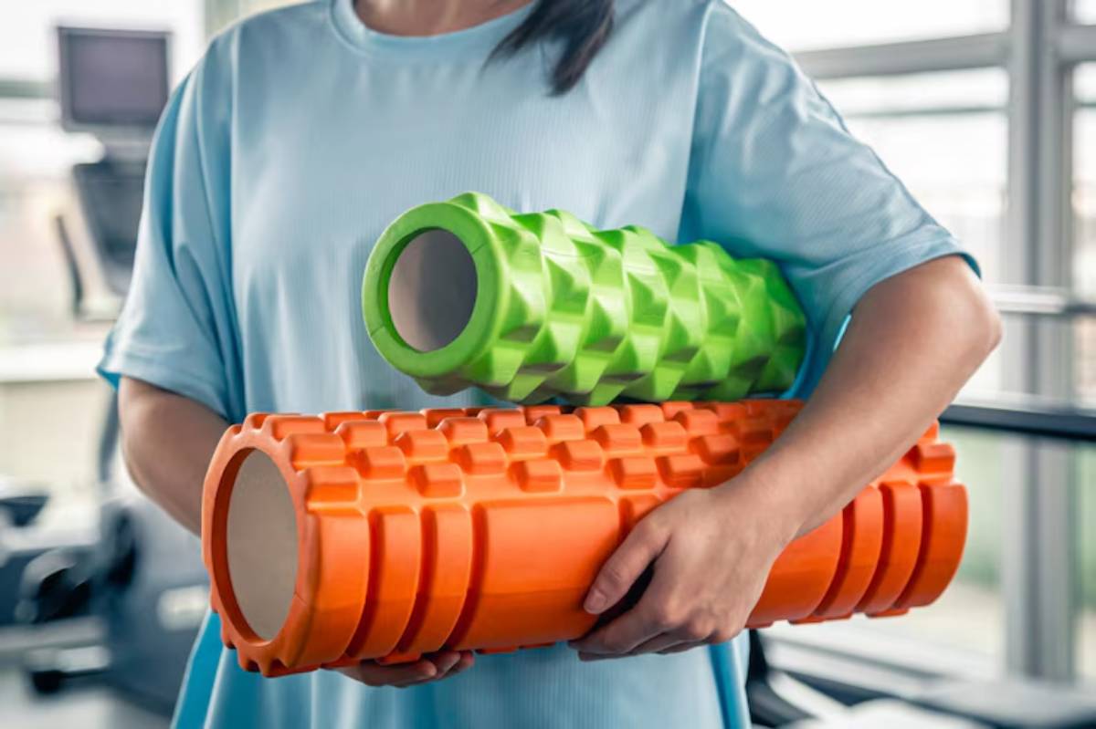 A person in a light blue shirt holds two foam rollers, one green with a textured surface and one orange with a ribbed design.