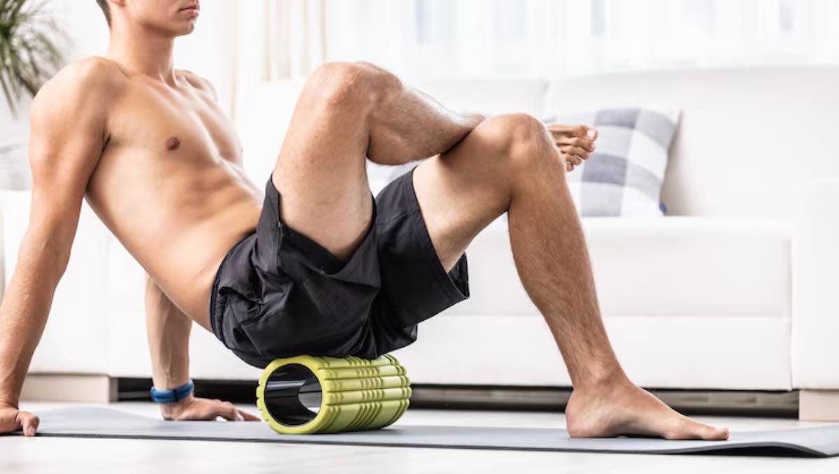 A young man rolls his back on a green foam roller while sitting on a yoga mat in a bright, modern living room.