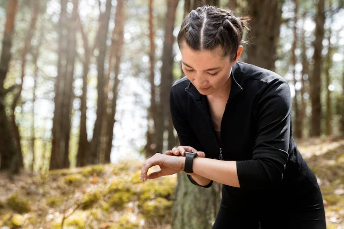 A person in a black athletic outfit checks their smartwatch while standing in a sunlit forest. Trees rise in the background.
