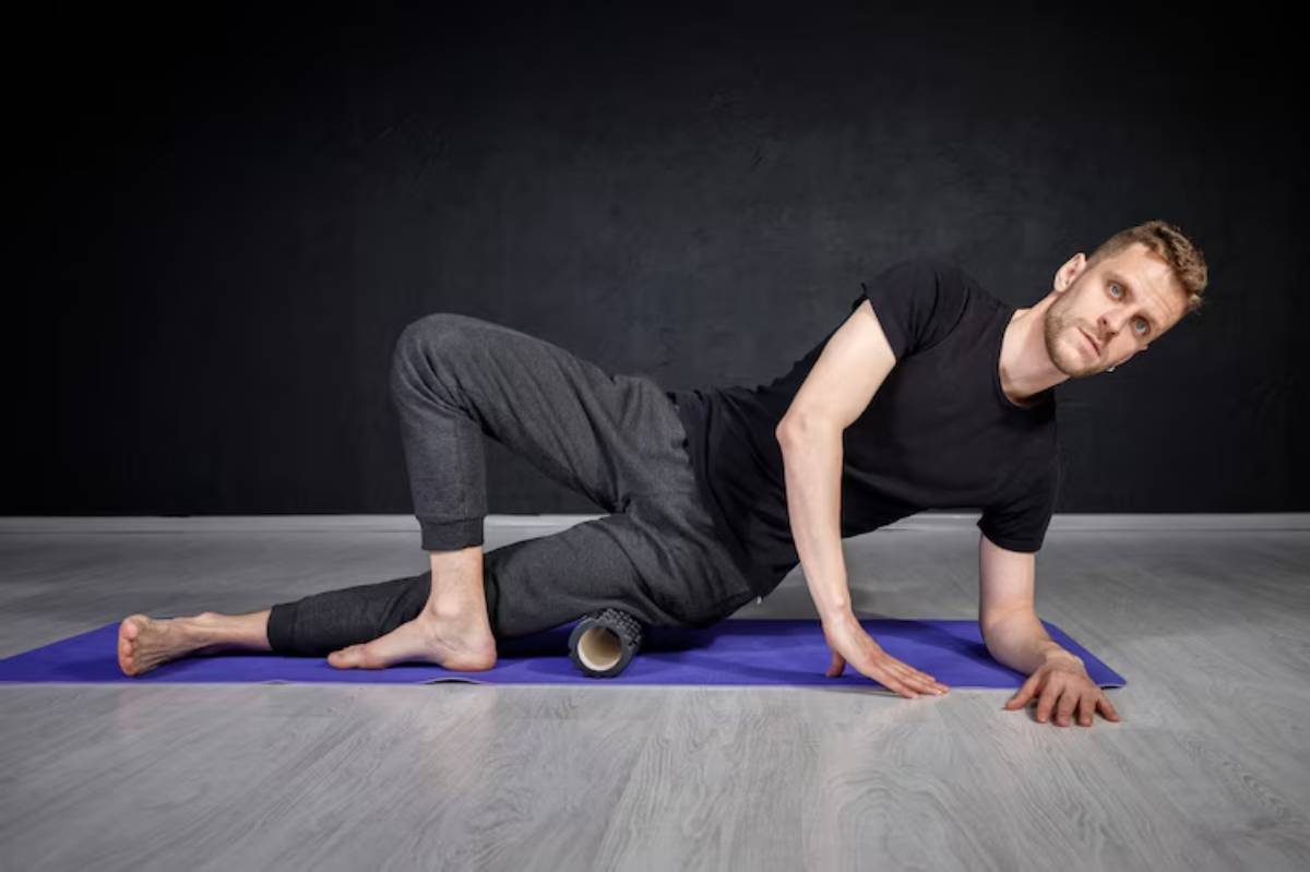 A person performs a side stretch on a yoga mat, supported by a foam roller, in a minimalist studio with dark walls.