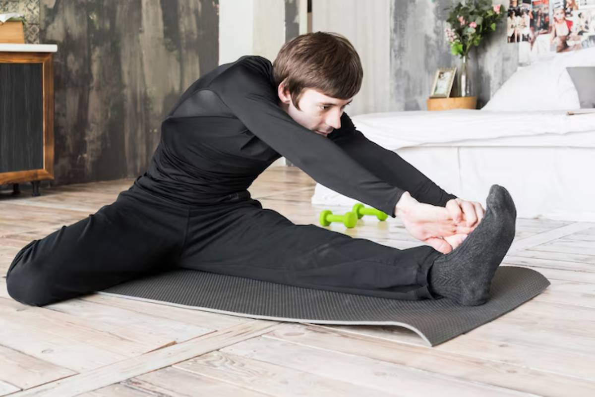 A person in black athletic wear stretches on a yoga mat in a bright, modern room, with green dumbbells nearby.