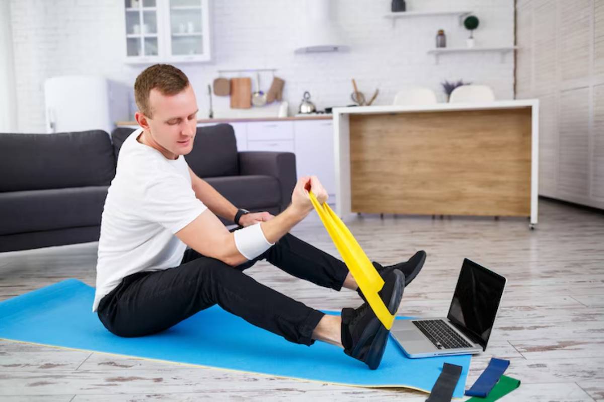 A person seated on a yoga mat at home performs resistance band exercises, with a laptop and fitness bands nearby.