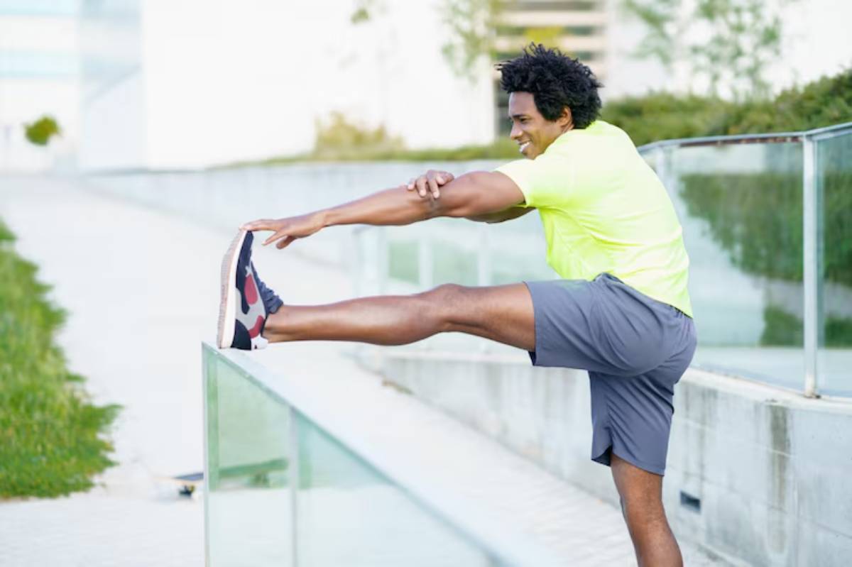 A person in a bright yellow shirt stretches their leg on a railing in an urban setting, preparing for exercise or running.