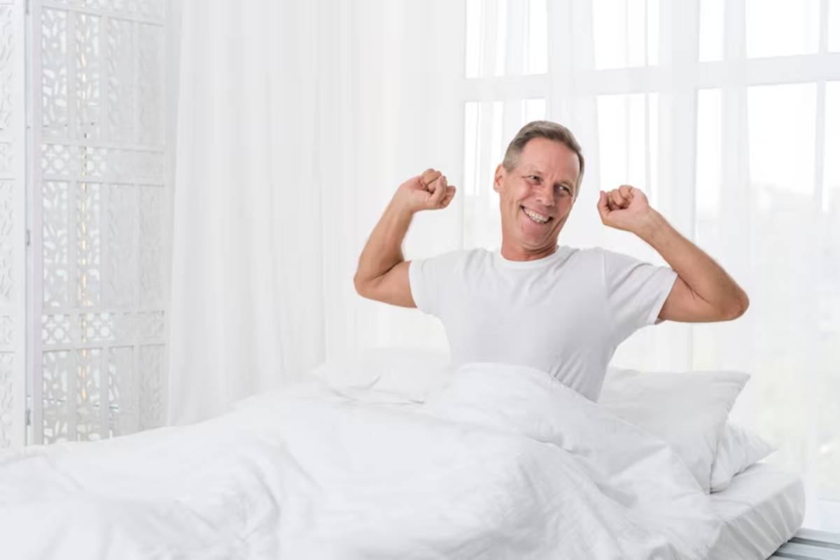 A man stretches in bed, wearing a white t-shirt, with white sheets and bright curtains filtering light into the room.
