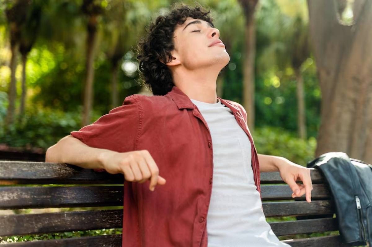 A person in a red shirt relaxes on a bench in a lush green park, enjoying the peaceful surroundings with closed eyes.