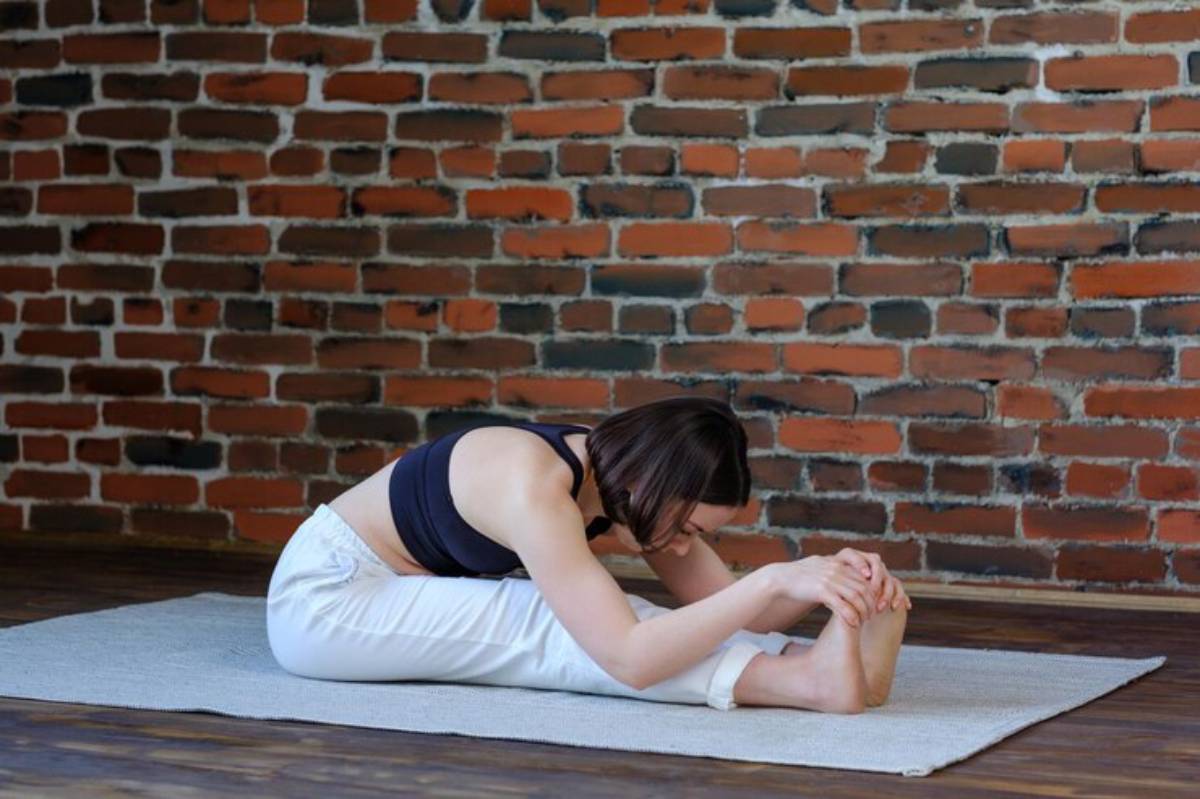 A woman in a black crop top and white pants stretches forward, reaching for her toes on a yoga mat against a brick wall.