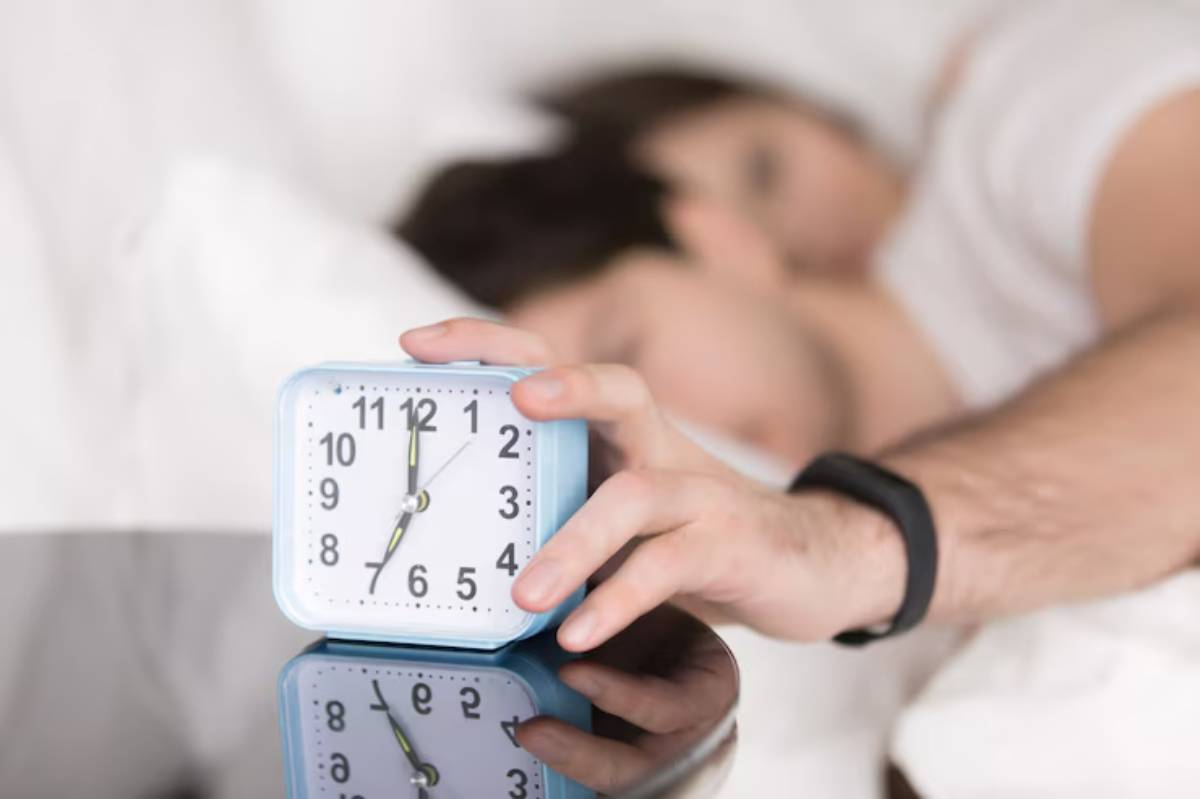 A person reaches for a blue alarm clock on a bedside table while lying in bed next to a partner, with soft bedding in the background.