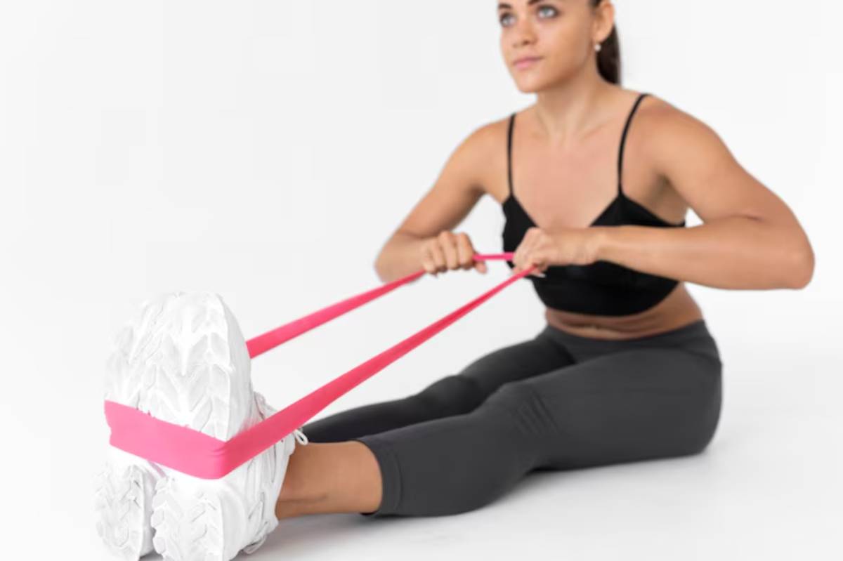 A person exercises using a pink resistance band while sitting with their legs extended in front, wearing a black tank top and leggings.