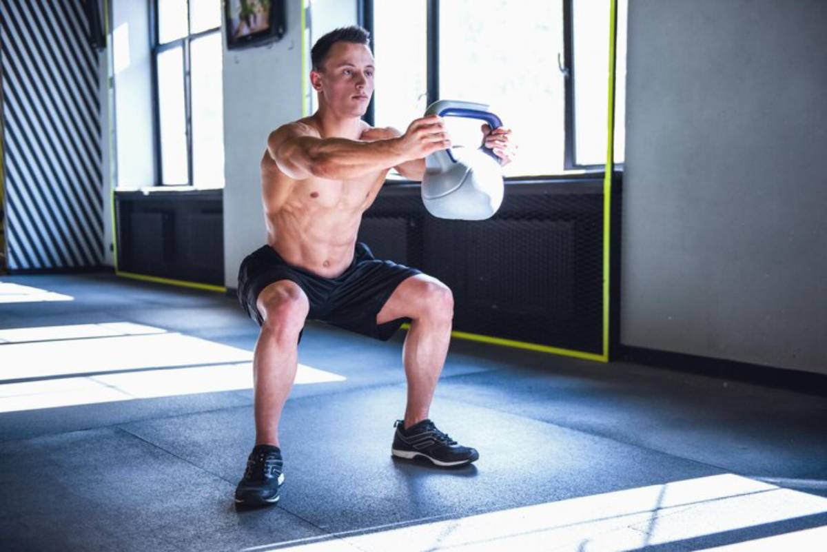A muscular man performs a squat while holding a kettlebell in a well-lit gym, showcasing strength and focus during his workout.