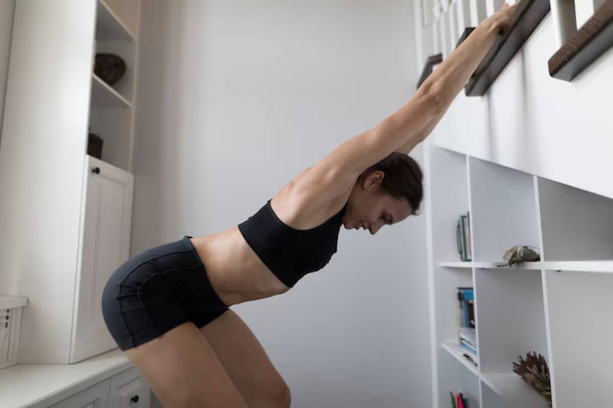 A person performs an exercise, leaning forward with arms extended towards a wall in a bright, modern room with white shelves.