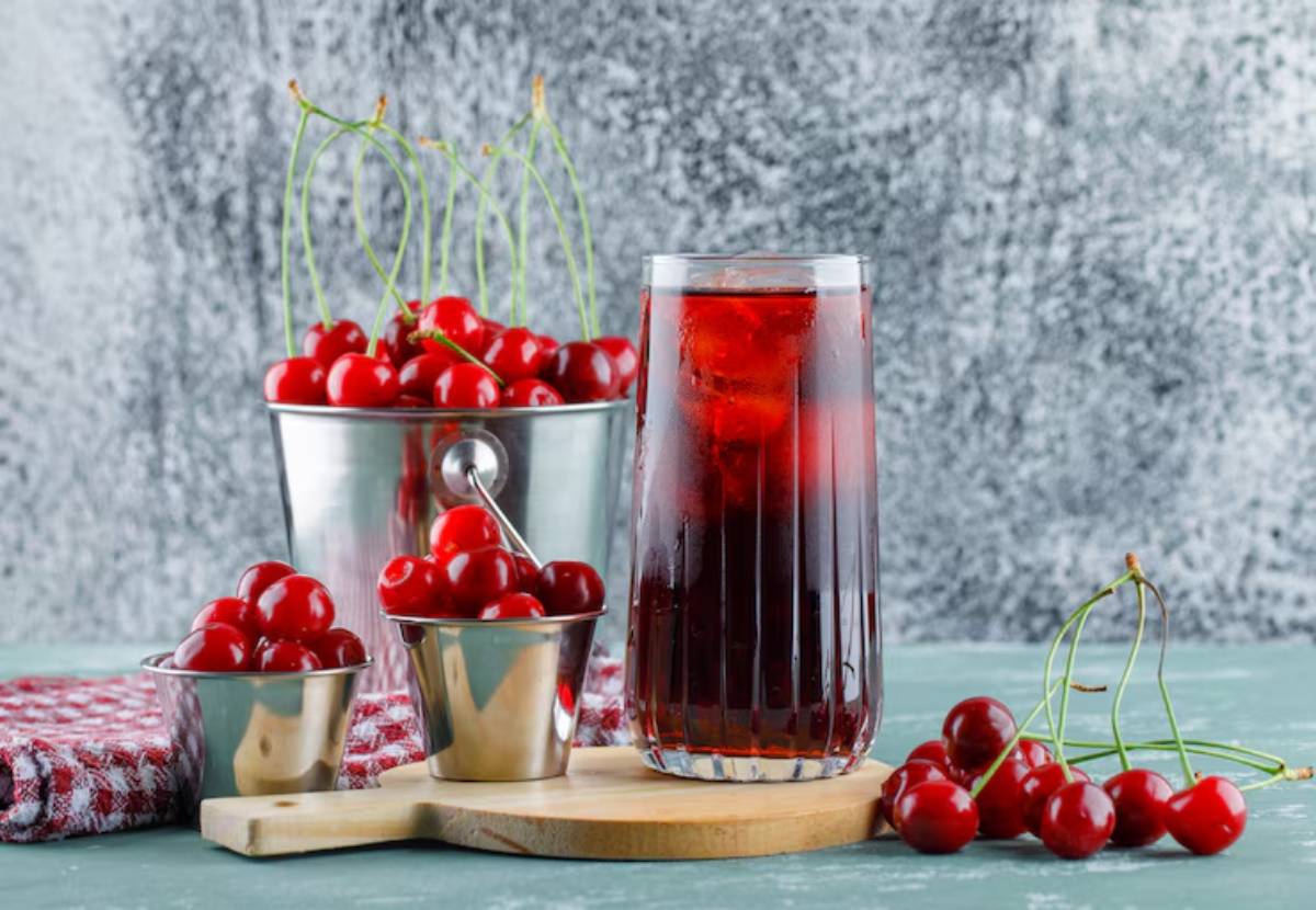 A tall glass of iced cherry drink surrounded by fresh cherries in metal buckets on a wooden board, with a textured gray background.
