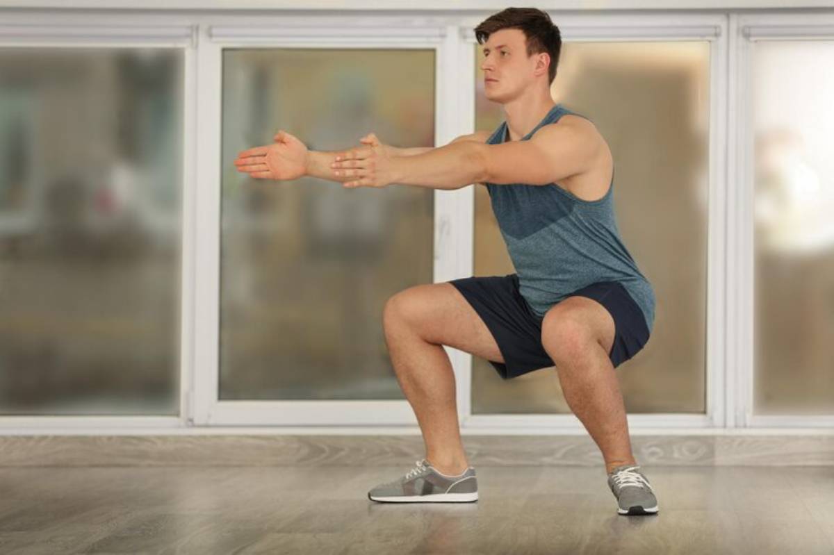 A man demonstrates a squat exercise in a bright room with large windows, wearing athletic gear and gray sneakers.