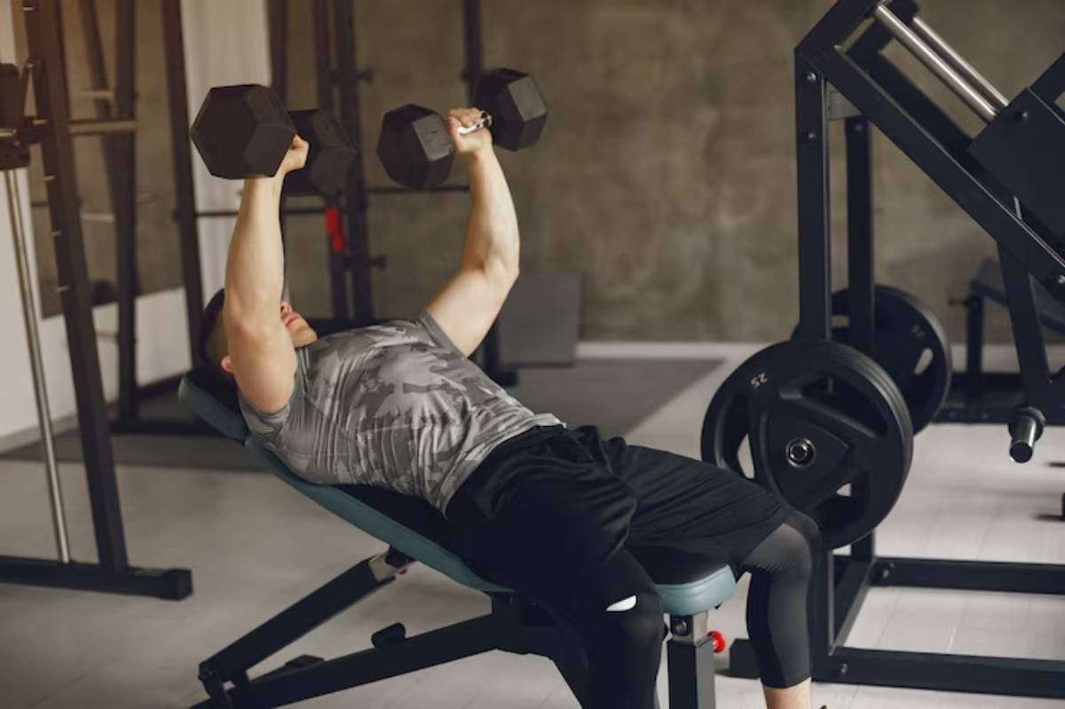 A man performs dumbbell bench press in a gym, lying on a bench with weights raised above his chest, focused on his workout.