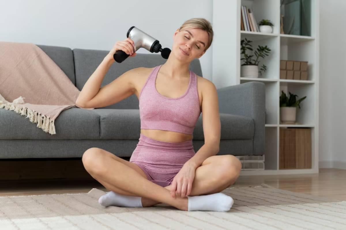 A woman sits cross-legged on a rug, using a massage gun while dressed in a pink workout outfit, with a cozy living room backdrop.