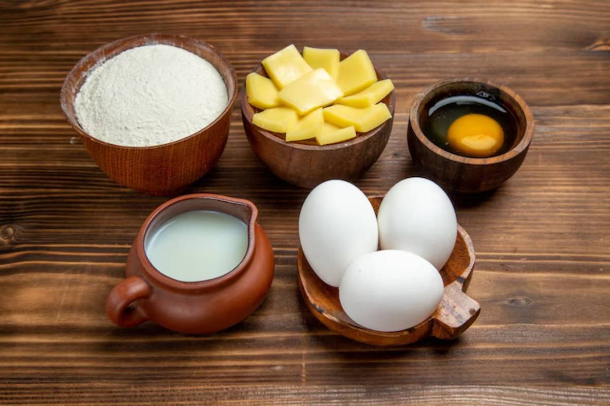 A collection of ingredients for cooking: flour, diced mango, milk, raw eggs, and three whole eggs arranged on a wooden surface.