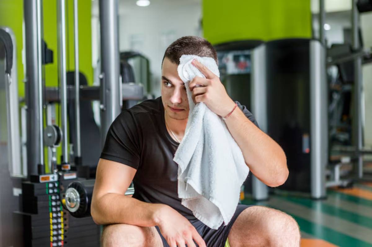 A man in a black workout shirt sits on the gym floor, wiping sweat from his forehead with a towel, surrounded by gym equipment.