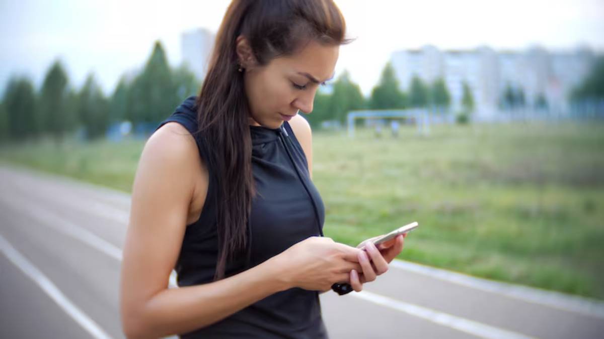 A woman in a sleeveless black top holds a smartphone while standing on a running track with a green field in the background.