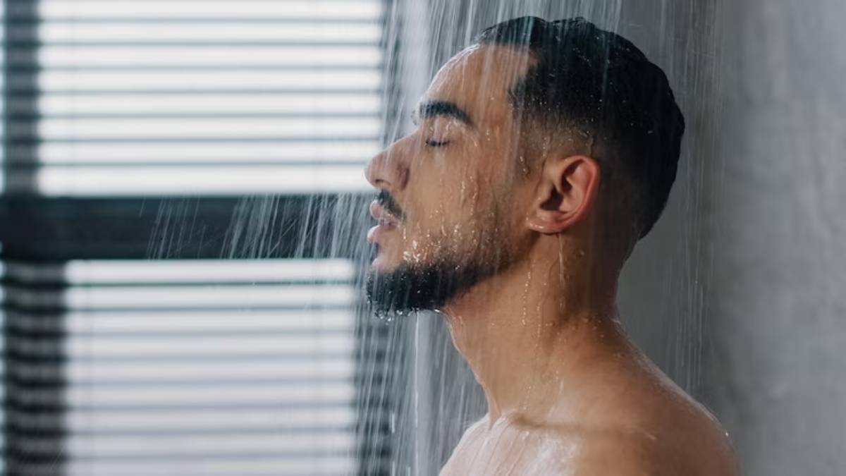 A person stands under a shower with water cascading down, surrounded by soft light filtering through window blinds.