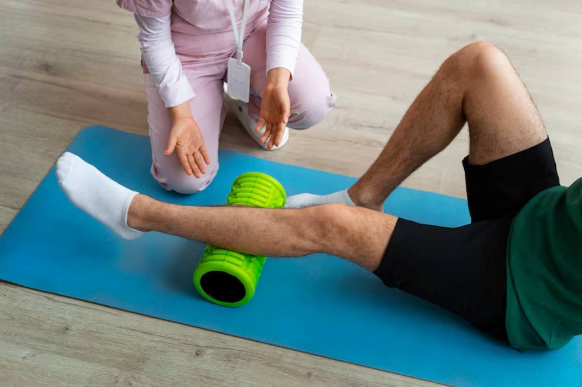 A therapist guides a patient using a green foam roller on a yoga mat for leg rehabilitation or stretching exercises.