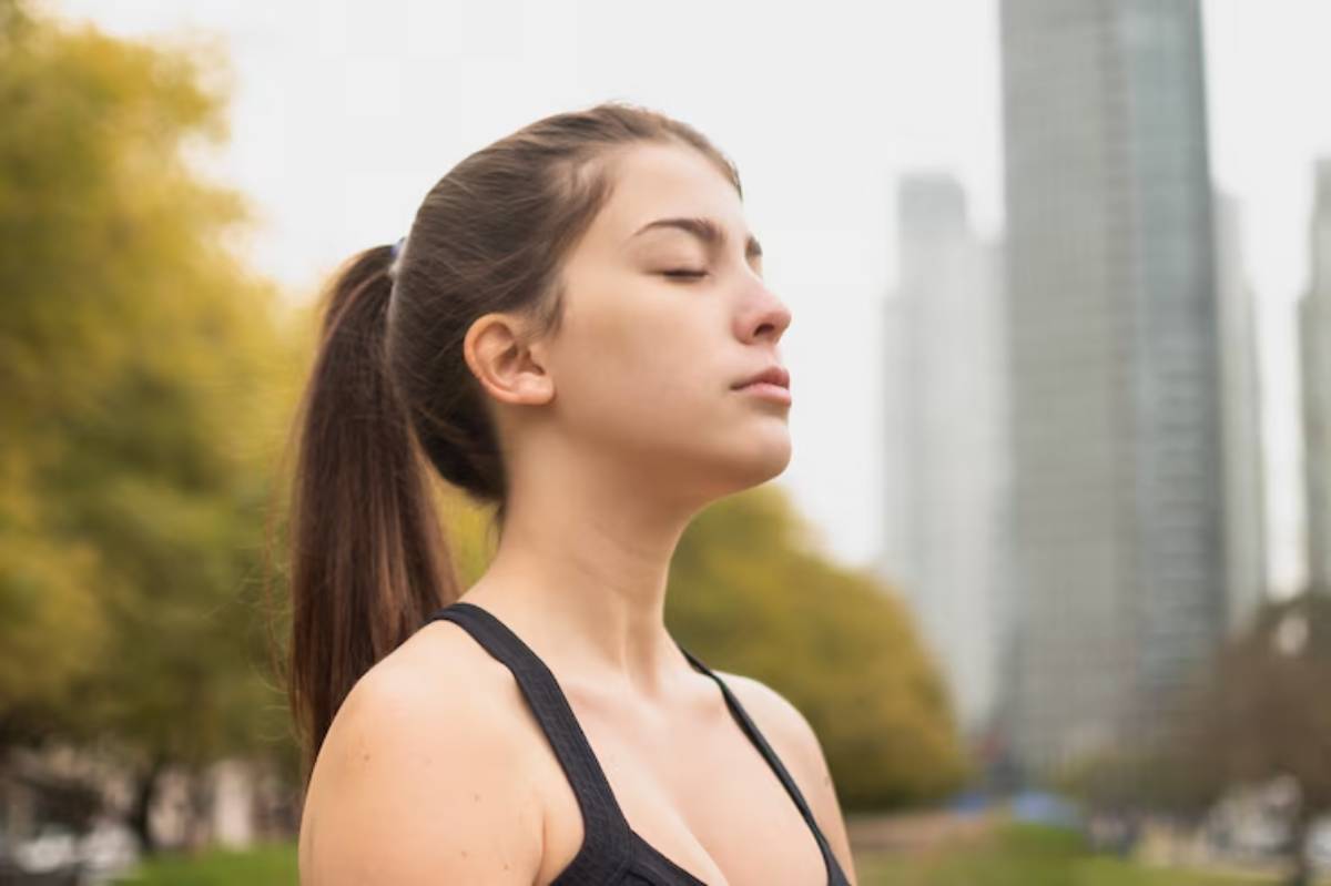 A woman with long hair tied in a ponytail stands outdoors, wearing a black sports top, with urban buildings and autumn trees in the background.