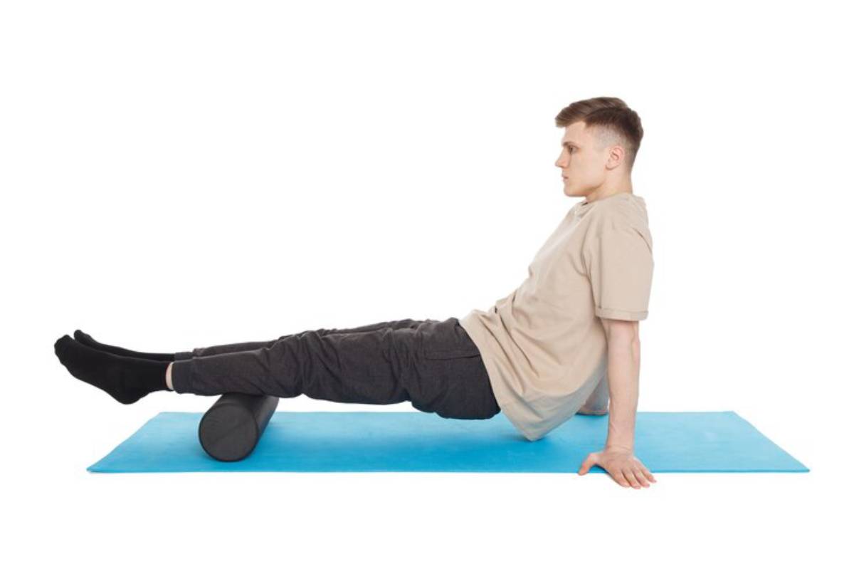 A person performs a stretching exercise on a blue mat, using a foam roller for support while seated with legs extended.