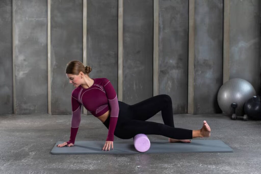 A woman practices yoga on a mat, using a pink foam roller for stretching and balance in a modern gym with gray walls.