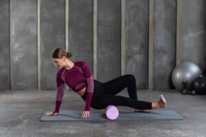 A woman practices yoga on a mat, using a pink foam roller for stretching and balance in a modern gym with gray walls.