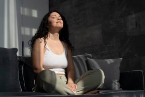 A person sits cross-legged on a couch in a sunlit room, wearing a tank top and light-colored pants, with dark textured walls in the background.