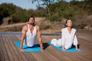 A man and a woman practice yoga on blue mats on a wooden deck, surrounded by greenery and a pool in a serene outdoor setting.