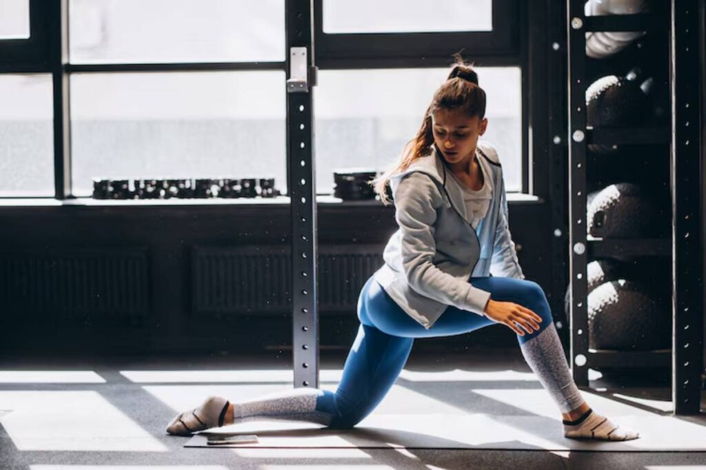 A person in activewear performs a lunging exercise on a yoga mat in a bright gym with weights in the background.