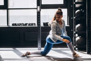 A person in activewear performs a lunging exercise on a yoga mat in a bright gym with weights in the background.