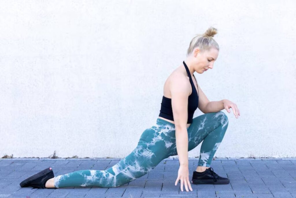 A person performs a lunging stretch outdoors, wearing a black top and patterned teal leggings, against a white wall.
