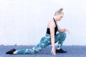 A person performs a lunging stretch outdoors, wearing a black top and patterned teal leggings, against a white wall.